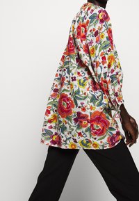 Hedvábná blůza s květinovým vzorem a mnohobarevným vyšíváním na světlém pozadí, volný střih, dlouhé puff rukávy a elastické manžety, kombinována s černými kalhotami.