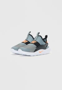 Un paio di sneakers slip-on Nike grigie e nere con dettagli arancioni e suole bianche, esposte in modo angolato su sfondo bianco.