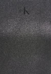 Tissu noir avec une texture scintillante, présentant un effet dégradé subtil et un logo CK en relief près du haut.