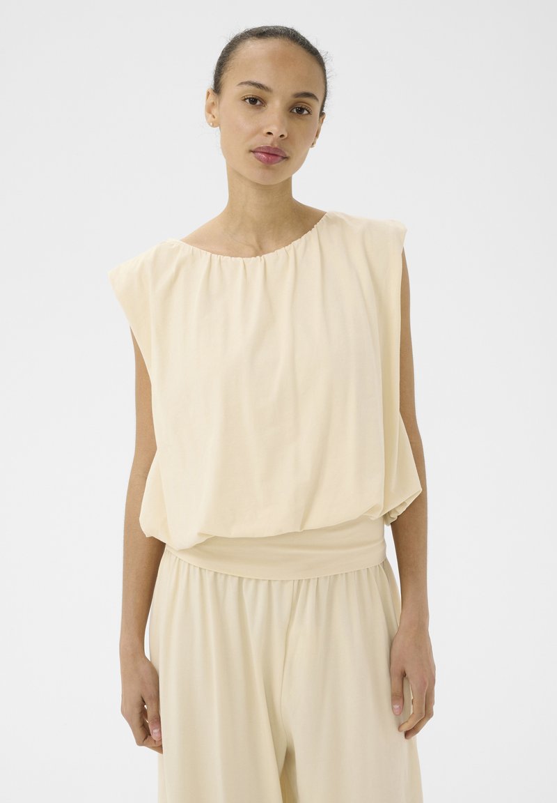 Jeune femme aux cheveux tirés en arrière, portant un haut beige ample sans manches et un pantalon large assorti, sur fond blanc.