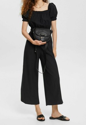 Zwarte leren crossbodytas met een afgeronde vorm, decoratieve klepsluiting en verstelbare riem. Model draagt een zwarte top en broek met wijde pijpen.