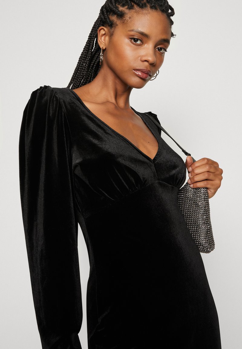Schwarzes Samtkleid mit tiefem V-Ausschnitt, Puffärmeln und taillierter Silhouette. Es hat eine Kettenriemen-Tasche in der rechten Hand des Modells.
