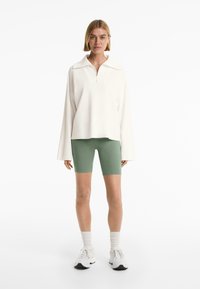 Pullover bianco a zip quarti con colletto ampio, abbinato a pantaloni ciclistici verde oliva. La modella indossa sneakers bianche e calzettoni bianchi a coste.