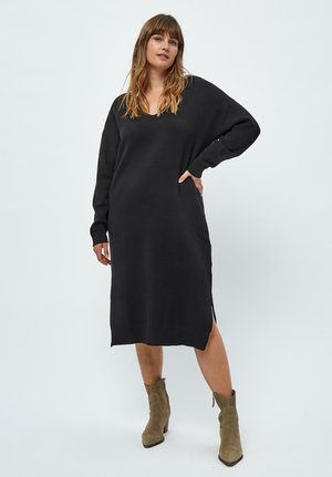 Black knitted dress with a V-neck, long sleeves, and side slits. The fabric appears soft with a slight texture. Styled with olive ankle boots.