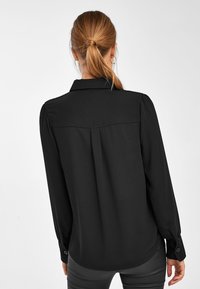 Blouse noire à manches longues avec col, présentant un pli dans le dos et des poignets boutonnés. Texture lisse ; coupe ajustée.