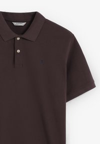 Polo shirt marrón oscuro en tejido texturizado, que presenta un cuello, una abertura de tres botones y un pequeño logo de calavera en azul marino en el lado izquierdo del pecho.