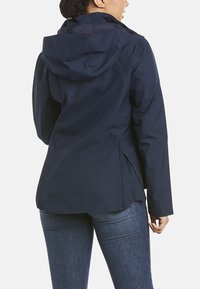 Veste imperméable navy avec capuche, poches latérales zippées et poignets élastiques, offrant une texture lisse et un design ajusté.