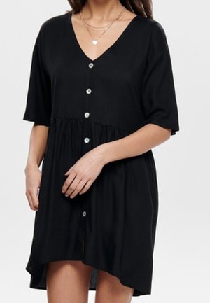 Kvinde iført en kortærmet sort kjole med knapper, V-hals og indsnævret talje, accessorized med lagdelte halskæder.