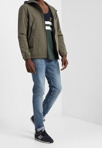 Man som bär en olivgrön huva jacka, färgblockerad marinblå och grön skjorta, ljusblå jeans och svarta New Balance sneakers, stående mot en vit bakgrund.