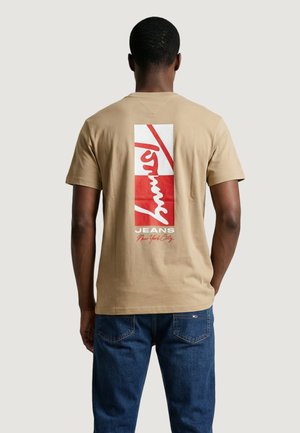 Uomo che indossa una t-shirt beige con grafica rettangolare rossa e bianca e testo "Tommy Jeans New York City" sul retro, abbinata a jeans blu.