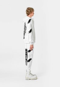 Sudadera con capucha blanca con patrones de alas negras, combinada con pantalones blancos que presentan diseños similares y detalles gráficos, complementados con botas blancas gruesas.