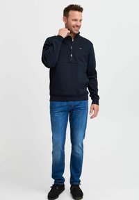 Sudadera negra con cremallera hecha de tejido suave, con un cuello de media cremallera, jeans azules y zapatos oscuros. Logo de la marca visible en el pecho.