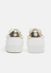 Fehér tornacipők texturált felülettel és arany színű fémes részletekkel a saroknál. Vastag, fehér gumitalppal és lekerekített orrkialakítással rendelkeznek.