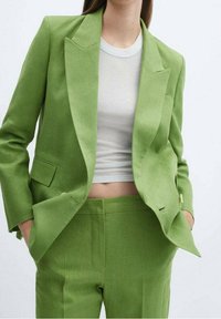 Costume deux pièces en lin vert comprenant une veste cintrée à revers crantés et un pantalon à taille plate, associé à un t-shirt gris clair.