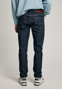 Jeans de mezclilla azul oscuro con un corte ajustado, que cuentan con un parche de cuero marrón en la parte trasera, costuras en contraste y bolsillos traseros estándar.