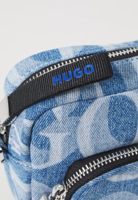 Borsa a tracolla in denim con un motivo astratto blu, dettagli in zip neri e un'etichetta con la scritta "HUGO" in lettere blu. Hardware in argento visibile.