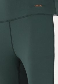 Leggings verde scuro realizzati in un materiale morbido e elastico, con cuciture nere e un piccolo dettaglio del logo argentato vicino alla vita.