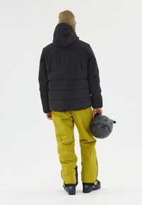 Svart puffjacka med huva, i kombination med ljusgula snöbyxor. Personen håller en grå skid hjälm. Släta texturer, funktionell design.