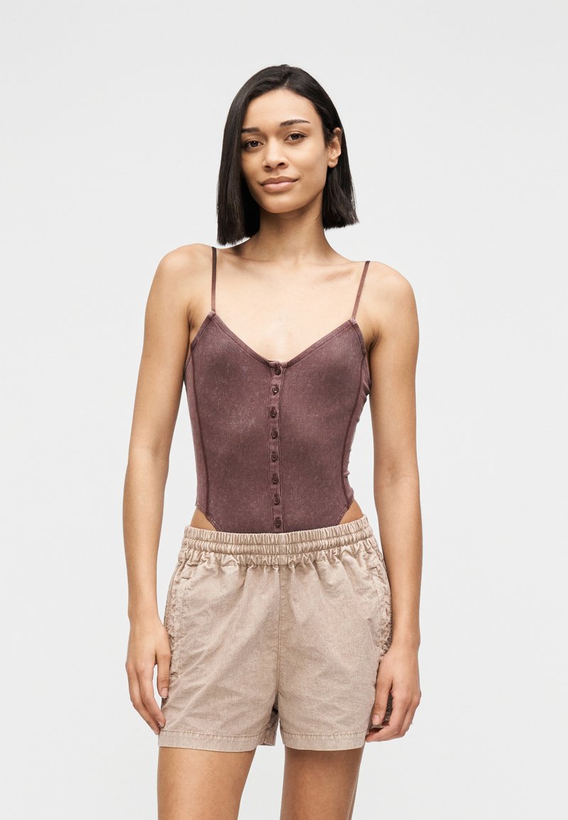 Νεαρή γυναίκα με ίσια σκούρα μαλλιά φοράει αμάνικο καφέ τοπ με κουμπιά και μπεζ σορτς με ελαστική μέση, σε απλό φόντο.