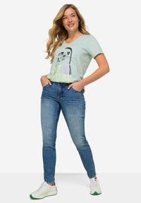 Ljust grön t-shirt med en sälgrafik och texten "GO STRAND", kombinerad med tajta blå jeans och vita sneakers med gröna inslag.