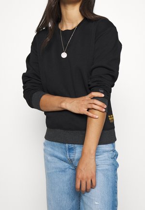 Žena v černé mikině s dlouhým rukávem a detaily na manžetách a v světle modrých džínách, držící levou předloktí pravou rukou.