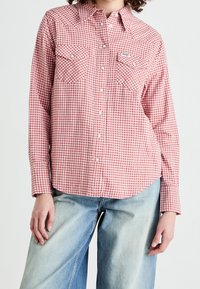 Camicia a quadri rossa e bianca con maniche lunghe, due tasche sul petto e design a yoke in stile western. Indossata con jeans larghi di colore blu chiaro.