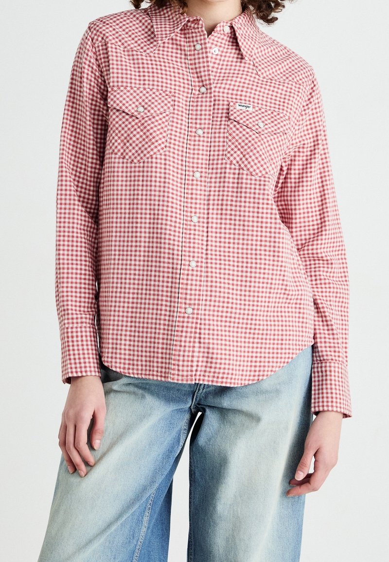 Camicia a quadri rossa e bianca con maniche lunghe, due tasche sul petto e design a yoke in stile western. Indossata con jeans larghi di colore blu chiaro.