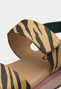 Sandale à bout ouvert avec semelle beige, brides rayées jaune et noir façon tigre, et bride arrière élastique verte, montrée en gros plan.