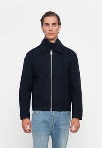 Man wearing a navy zip-up jacket with collar over a dark turtleneck sweater and light blue jeans, standing against a plain white background.