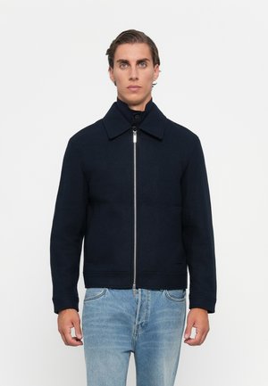 Mann iført en marineblå jakke med glidelås og krage over en mørk høyhalset genser og lyseblå jeans, stående mot en enkel hvit bakgrunn.