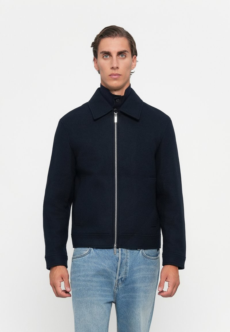 Man wearing a navy zip-up jacket with collar over a dark turtleneck sweater and light blue jeans, standing against a plain white background.