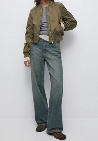 Mujer vestida con una chaqueta bomber verde oliva, suéteres en capas gris y crema, jeans de campana azules y botas marrones de punta cuadrada, de pie contra un fondo blanco.