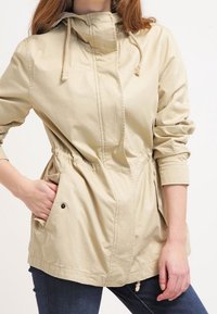 Ljust beige jacka med framdragkedja, dragsko huva, elastisk midja och två sidofickor. Tillverkad av bomull med en mjuk yta.