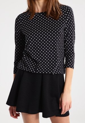 Kobieta ubrana w czarną bluzkę z długim rękawem w białe groszki i czarną rozkloszowaną spódnicę, stojąca na tle jednolitego tła.