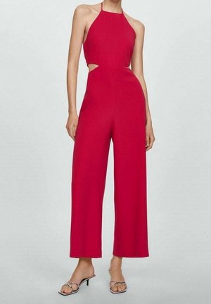 Femme portant une combinaison sans manches rouge à col halter, jambes larges et découpes à la taille, associée à des sandales à talons ouvertes argentées.