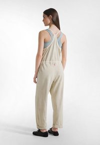 Béžový overal s volným střihem, s designem na zádech ve tvaru závodnických pásků a elastickým pasem. Kombinován se světle modrým tílkem a černými botami.