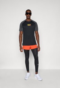 Sort atletisk t-shirt med grå ærmer, "BERLIN RUN" trykt i gul, parret med orange shorts og sorte leggings. Hvide sneakers fuldender looket.