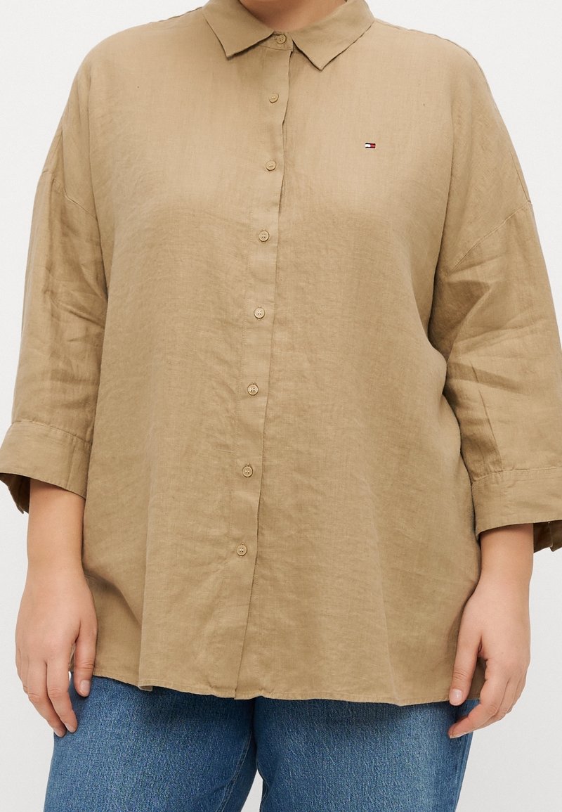 Camisa de lino beige con cuello, botones en la parte delantera y mangas de tres cuartos. Presenta un pequeño logo bordado en el pecho.