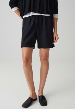 Persoon draagt zwarte shorts met trekkoord, zwarte instapschoenen en gelaagde zwart-witte tops met lange mouwen, staand tegen een effen achtergrond.