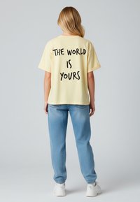 T-shirt in cotone giallo con una vestibilità rilassata, caratterizzato da un grande testo nero "IL MONDO È TUO" sul retro; indossato con pantaloni jogger azzurri chiari.