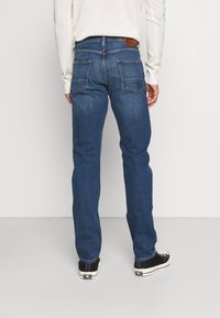 Man wearing blue straight-leg jeans, white long-sleeve shirt, and black low-top sneakers, standing against a plain white background.