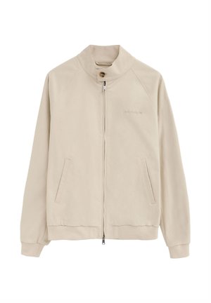 Chaqueta beige con cremallera, cuello alto con botón, mangas largas, bolsillos laterales y un logo de marca sutil en el pecho.
