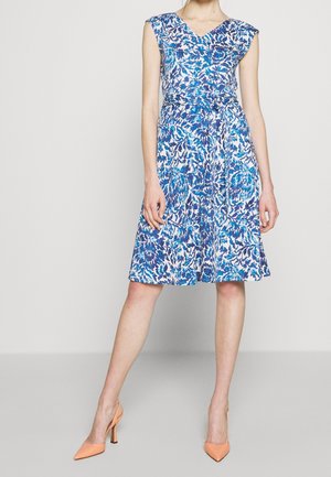 Femme portant une robe sans manches bleue et blanche à fleurs, longueur genoux, avec un col en V et des chaussures à talons hauts pêche, debout devant un fond uni.