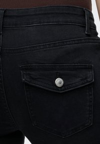 Gros plan de la poche arrière d’un jean en denim noir avec rabat et fermeture par bouton-pression argenté.