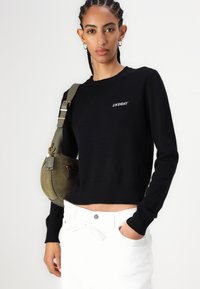 Sort strikket sweater med rund hals og ribbekantede ærmer, der har hvid "2NDAY" broderi. Sammen med hvide bukser og en olivenfarvet skuldertaske.