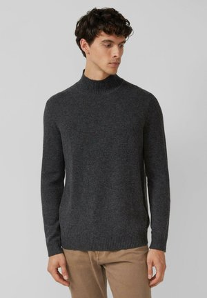 Homme portant un pull col roulé gris foncé et un pantalon beige, regardant sur le côté devant un fond clair.
