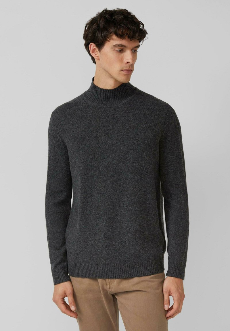 Uomo che indossa un maglione a collo alto grigio scuro e pantaloni beige, che guarda di lato su uno sfondo chiaro e uniforme.