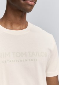 Gaiši beigu īsām piedurknēm T-krekls, izgatavots no mīksta kokvilnas, ar uzrakstu "Denim Tom Tailor Established 2007" drukātu maigi zaļā krāsā.