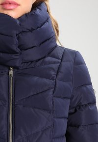 Marinblå puffjacka med hög krage, med kviltad textur, frontdragkedja och diskreta metallisk detaljer.