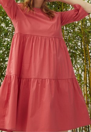 Femme en robe corail ample à étages avec manches jusqu'aux coudes, se tenant parmi de hauts bambous verts, les bras levés près de la tête.